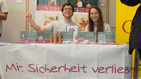 Zwei Studierende sitzen hinter einem Tisch, auf dem zahlreiche Flyer, aber auch Präparate für die Unterrichtseinheiten liegen. Auf der Tischdecke steht: "Mit Sicherheit verliebt - Lokalgruppe Mainz".