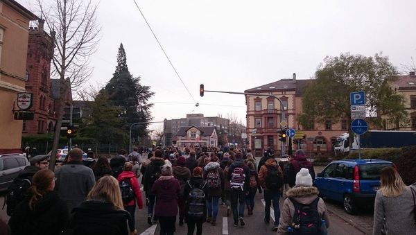 Ein Demonstrationszug zieht durch die Straßen. Auf der Straße sieht man viele Menschen, links und rechts sowie im Hintergrund Autos und Häuser. Die Menschen überqueren eine große Kreuzung mit Ampel. 