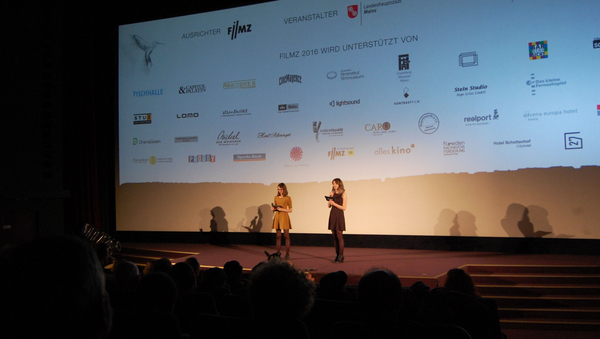 Zwei Frauen auf einer Bühne vor einer großen Leinwand. 