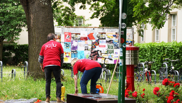 Links steht ein Mann und rechts eine Frau in gebückter Haltung, beide in roten T-Shirts und mit dem Rücken zur Kamera, vor einer Wiese. Im Hintergrund steht ein großer Baum, rechts daneben eine Plakatwand, ein roter Hydrant und rote Rosen.