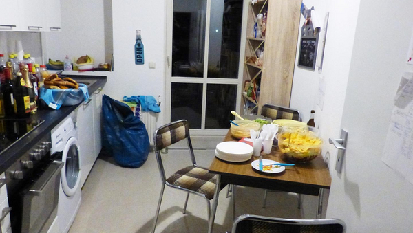 Blick in eine Küche. Links Backofen, Waschmaschine, Arbeitsfläche, Schränke, etc. Rechts an der Wand ein Tisch und Stühle. Auf dem Tisch steht eine Schüssel mit Taco-Salat. Im Hintergrund, am Ende des Raumes, ein Schrank und eine Glastür. 