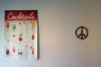 Eine weiße Wand. Links ein Poster, auf dem verschiedene Cocktails abgebildet sind. Rechts Bierkronen, die in Form eines Peace-Zeichens aufgehängt sind. 