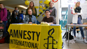 Vier Studierende sitzen hinter einem Tisch, welcher mit dem Logo von Amnesty International verkleidet wurde.